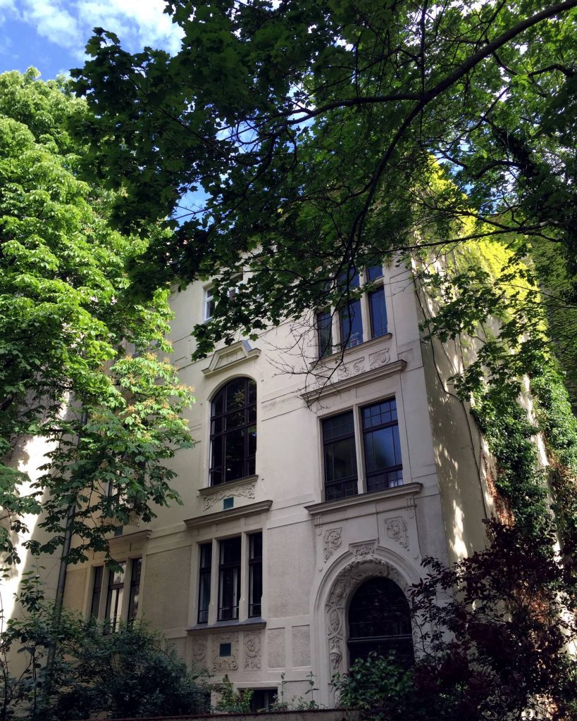 Die Stuckfassade der Alten Artistenschule zwischen Bäumen: hohes Bogenfenster, darunter ein ornamentiertes Rundbogenportal.